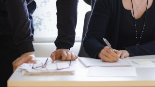 Pöydällä on papereita ja silmälasit, yhden henkilön kädet nojaavat pöytää vasten ja toinen henkilö kirjoittaa paperille. 