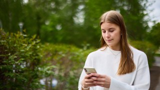 Nuori nainen katsoo puhelintaan. Taustalla puita ja pensaita.