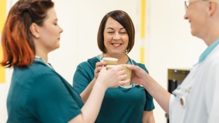 Kaksi hoitajaa ja lääkäri kippistävät pahvimukeilla.