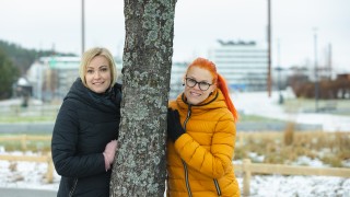 Kaksi naista nojaa puuhun eri puolilta alkukeväisessä kaupunkimaisemassa.