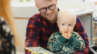 Lapsi istuu isän sylissä, isä hymyilee ja lukee lapselle kuvakirjaa.