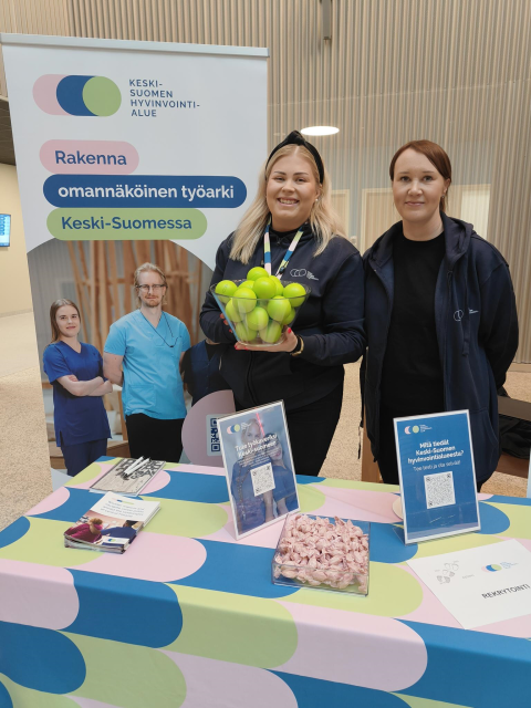 Rekrytointipalveluiden kaksi asiantuntijaa hymyilee esittelypisteen takana. Toisella on kädessä kulhollinen kirkkaan värisiä palloja. 